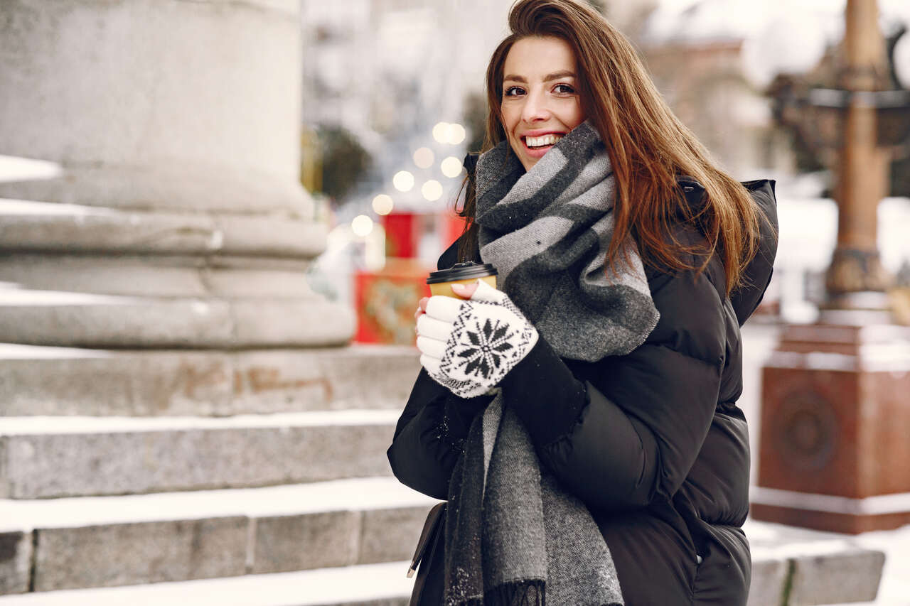 Smiling woman holding coffee in Budapest during winter, wearing a black puffer jacket, gloves, and scarf — practical outfit for cold weather.