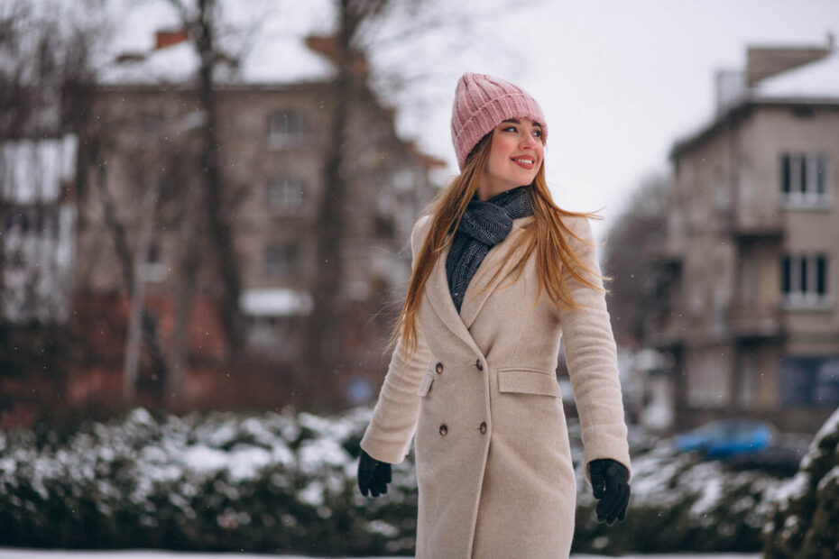 Young woman in beige wool coat and pink beanie walking in snowy Budapest — stylish and warm winter outfit idea.