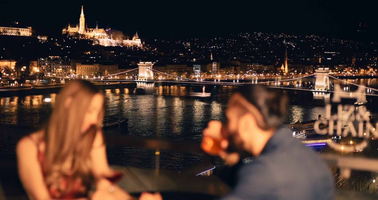 Couple enjoying drinks at a rooftop bar overlooking the Danube and the illuminated Chain Bridge at night, symbolizing a romantic evening in Budapest.