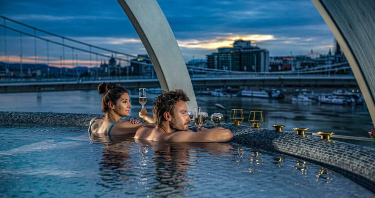 Couple relaxing in an outdoor thermal bath at night with the Danube and Budapest skyline in the background, symbolizing a romantic thermal bath experience.