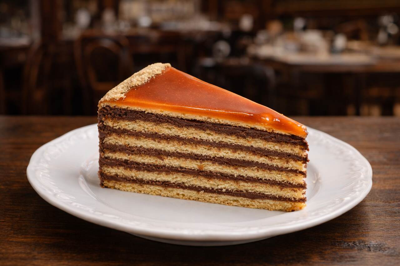Slice of Hungarian Dobos torte with multiple sponge and chocolate layers topped with caramel, served on a white dessert plate